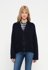 Granatowy sweter kardigan z dekoltem w serek, przodem na siedem guzików i dwiema kieszeniami z przodu. Miękka dzianinowa faktura i obniżone ramiona.