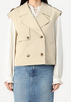 Frau trägt beige ärmellose, zweireihige Weste über weißer langärmeliger gestreifter Bluse und blauem Jeansrock, steht vor weißem Hintergrund.