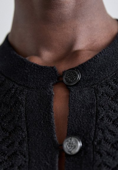 Sort tekstureret stof beklædning med rund halsudskæring, der har to fremtrædende dekorative knapper ved den åbne front. Diskret åbenværksmønster.