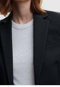 Zwarte blazer met gestructureerde revers, gecombineerd met een lichtgrijs geribbeld sweater met subtiele spikkels. Strak, modern ontwerp.