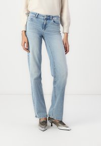 Jeans de mezclilla azul claro con un ajuste relajado, cinco bolsillos y un acabado ligeramente desgastado. Combinados con zapatos de tono plateado y una blusa de color claro.