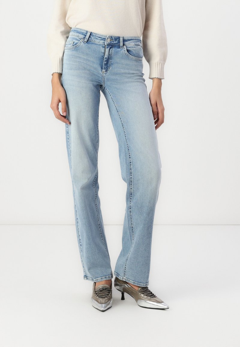 Jeans de mezclilla azul claro con un ajuste relajado, cinco bolsillos y un acabado ligeramente desgastado. Combinados con zapatos de tono plateado y una blusa de color claro.