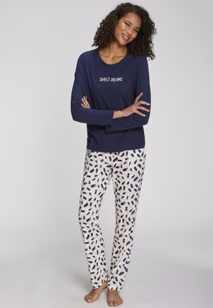 Frau mit lockigem Haar, die ein marineblaues langärmeliges Oberteil mit dem Schriftzug „SWEET DREAMS“ und weiße gemusterte Schlafanzughosen trägt, steht barfuß und lächelt.