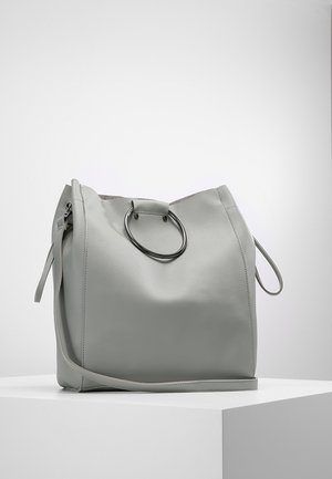 Lichtgrijze leren handtas met een groot cirkelvormig metalen handvat en een lange schouderriem, rustend op een wit voetstuk.