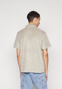 Khaki poloko s krátkým rukávem, vyrobené z měkké látky. Má límeček a volný střih. Na sobě má světle modré džíny, na rukávu je vidět tetování.