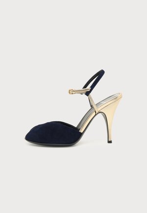 Escarpin bleu marine à bout pointu avec bride de cheville dorée et talon, vue de côté sur fond gris clair.