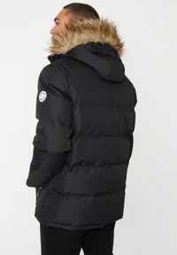 Czarna kurtka puchowa z kapturem wykończonym sztucznym futrem, poziomym pikowaniem i zamkami błyskawicznymi w kieszeniach, z okrągłym logo na rękawie.