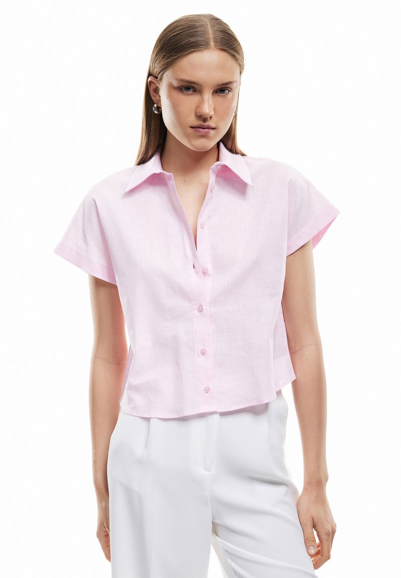 Joven mujer con cabello liso lleva puesta una camisa de botones de manga corta color rosa claro y pantalones blancos de talle alto sobre un fondo blanco.