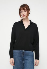 Young woman with red hair wearing a black long-sleeve top with a V-neck collar and blue jeans against a plain white background.