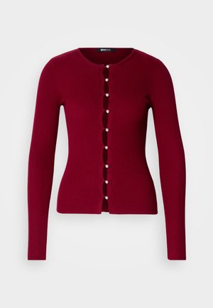Rdeča rebrasta kardigan s okroglo ovratnikom, ki ima vrsto bisernih gumbov in izrezane detajle spredaj. Dolgi rokavi.
