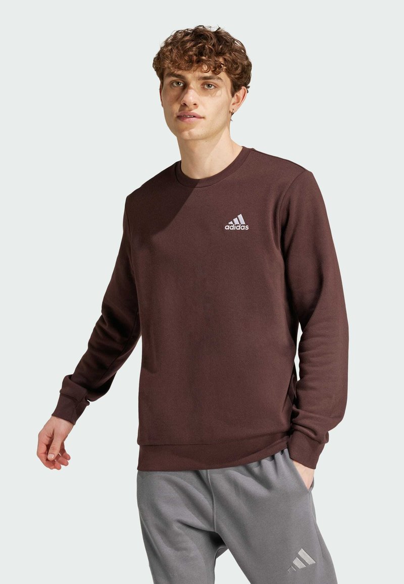 Brun sweatshirt med rund hals, långa ärmar och ribbade muddar. Har en vit Adidas-logotyp på bröstet. Matchas med grå byxor.