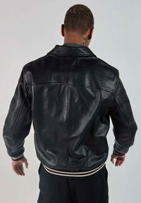 Veste en cuir noir avec une coupe décontractée, col cranté, poignets et ourlet côtelés, présentant une texture lisse et un design minimaliste.