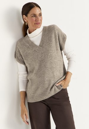 Vrouw in beige gebreid vest over een witte coltrui en donkere broek, staand met één hand in de zak, zijwaarts kijkend en glimlachend.