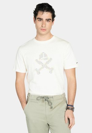 Joven con cabello oscuro que lleva una camiseta blanca con un diseño de bates cruzados y casco, pantalones gris claro y muñequeras en la muñeca izquierda.