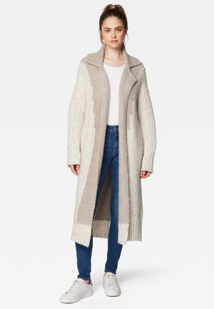 Langer, offener Cardigan in Creme und Beige. Texturierter Strick mit gerippten Ärmeln und einem breiten Kragen. Kombiniert mit blauen Jeans und weißen Turnschuhen.
