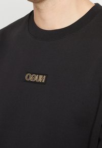 Sudadera negra con cuello redondo acanalado, que presenta una etiqueta rectangular negra con el texto dorado "HUGO" en la parte izquierda del pecho.