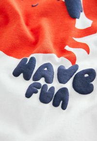 T-shirt en coton présentant un design graphique : soleil orange et texte "AMUSEZ-VOUS" en bleu marine avec une finition douce et texturée sur un fond blanc.