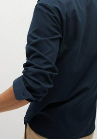 Uomo che indossa una camicia a maniche lunghe blu navy con le maniche arrotolate, abbinata a pantaloni beige, mostrato di spalle e di lato.