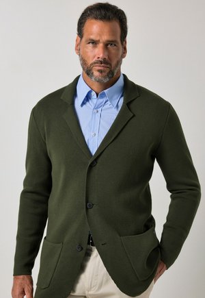 Man with beard wearing dark green blazer over light blue dress shirt and beige pants, standing with one hand in pocket against plain background.