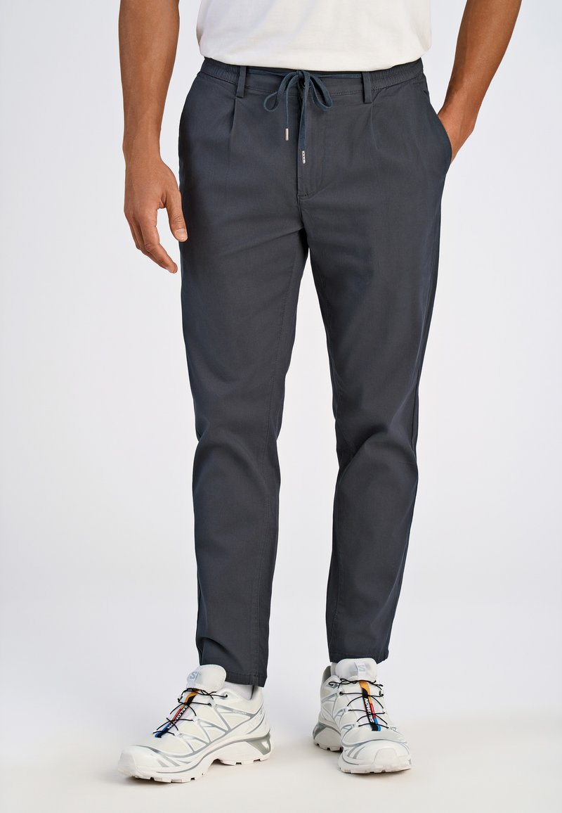 Pantalons légers gris foncé avec une taille à cordon, jambe fuselée, poches avant et texture lisse, associés à des chaussures de sport blanches.