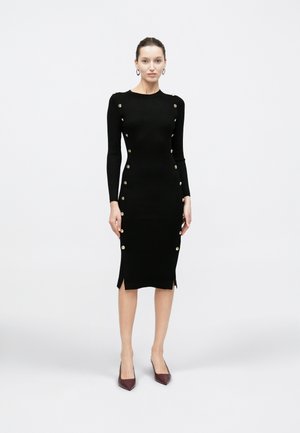 Vrouw staand in een zwarte geribbelde midijurk met gouden knopen aan de zijkanten en lange mouwen, draagt bordeauxrode puntige hakken.