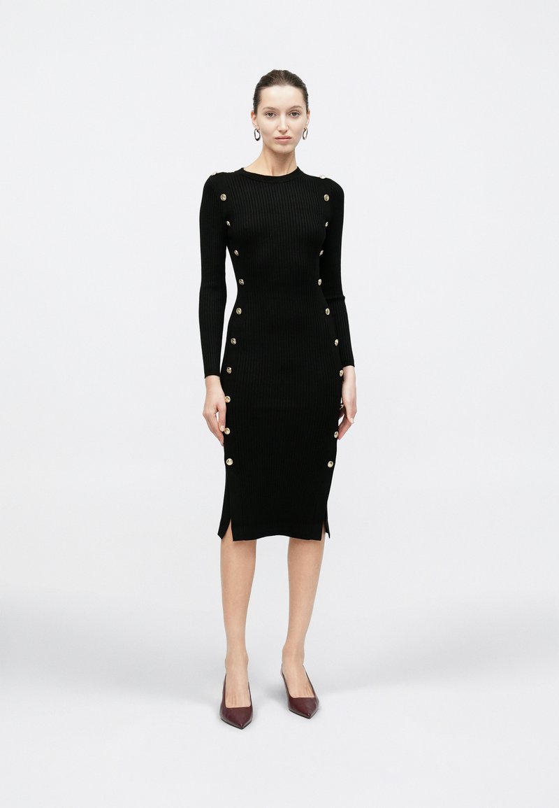 Kobieta stojąca w czarnej prążkowanej sukience midi z złotymi guzikami po bokach i długimi rękawami, ubrana w bordowe szpiczaste szpilki.