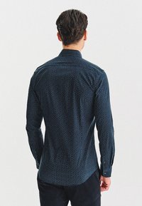 Mężczyzna stoi tyłem, ubrany w ciemnoniebieską koszulę z długim rękawem w mały biały wzór oraz czarne spodnie.