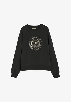 Czarny sweter z rękawami raglanowymi, z okrągłym złotym emblematem na przodzie, na którym widnieje napis "Różnorodne Dziedzictwo" w otoczeniu laurowego ornamentu.