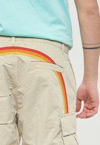 Pantaloni cargo beige con un design a righe arcobaleno multicolori in rosso, arancione e giallo sulla tasca posteriore. Tessuto morbido e resistente.