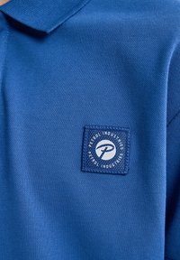 Modrá polo košile se strukturovanou tkaninou, se čtvercovým logem v bílé a námořnické barvě. S límečkem a upraveným designem.