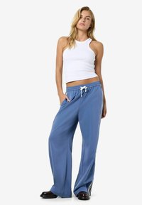 Pantalons larges bleu clair avec taille élastique, comportant des rayures blanches sur les côtés et deux poches latérales. Texture douce et fluide.