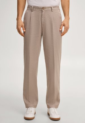 Man met beige geplooide broek en witte sneakers, staand met ontspannen handen langs het lichaam tegen een effen achtergrond.