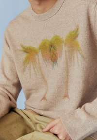 Pull beige à texture douce avec une impression abstraite colorée de feuilles de palmier. Encolure arrondie, poignets côtelés et coupe décontractée.
