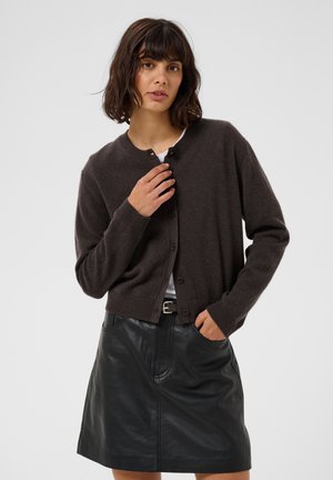Femme aux cheveux bruns foncés mi-longs, portant un cardigan marron foncé boutonné et une jupe en cuir noire, une main près du col et l'autre dans la poche.