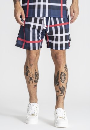 Mężczyzna w granatowo-biało-czerwonych kraciastych szortach z białym sznurkiem, białe trampki i czarne tatuaże na obu nogach stoi na białej podłodze.