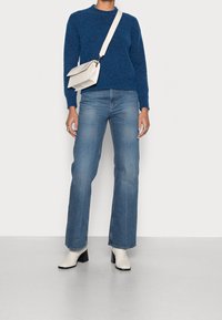 Pull bleu, jean bleu clair à jambe droite et bottines blanches à talons. Sac avec tissu texturé et bandoulière ajustable. Design minimaliste.