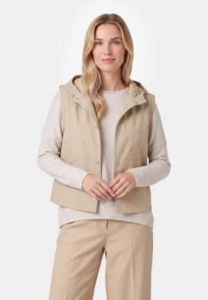 Donna bionda con capelli sciolti indossa un gilet beige senza maniche con cappuccio sopra una maglietta leggera a maniche lunghe con motivi e pantaloni beige, in piedi con le mani giunte.