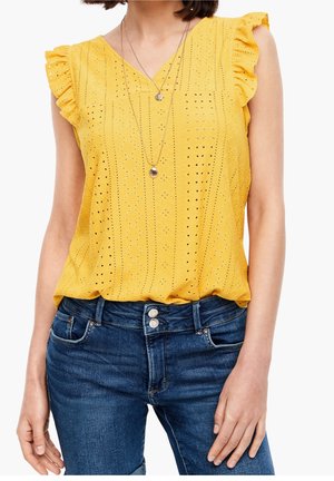 Kvinde iført gul ærmeløs top med hulmønster og flæser på skuldrene samt lagdelte sølvskalskæder, kombineret med blå jeans.