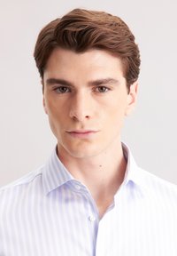 Jasnoniebieska koszula w paski z guziczkami i kołnierzykiem. Gładka tekstura materiału i dopasowany krój. Twarz modela pokazana, z neutralnym tłem.