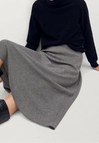 Persona sentada con una blusa negra de manga larga, una falda midi a cuadros en blanco y gris, y botas de tobillo negras, contra un fondo claro y sencillo.