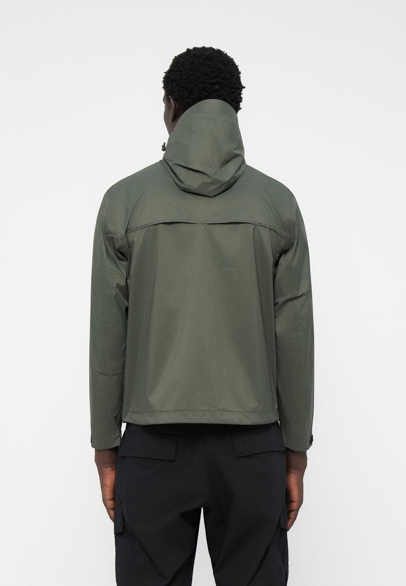 Homme portant une veste à capuche vert olive et un pantalon cargo noir, vu de dos sur un fond blanc uni.