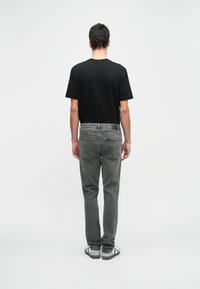 Homem de costas, vestindo uma camiseta preta, calças cinzentas e ténis brancos, contra um fundo branco simples.