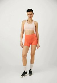 Reggiseno sportivo rosa chiaro e pantaloni corti alta vita arancioni brillanti, abbinati a scarpe da ginnastica nere e calzini bianchi a crew. Texture lisce e aderenti.