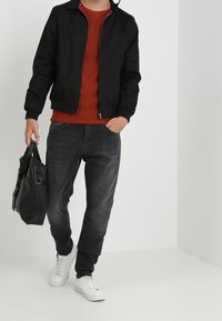 Czarna kurtka typu bomber na rdzawobrązowym swetrze, zestawiona z ciemnymi dżinsami i białymi trampkami. Nosi czarną skórzaną torbę.