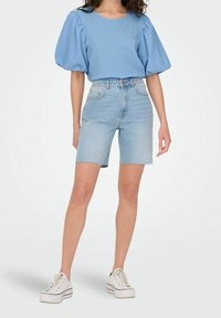 Haut à manches bouffantes texturé bleu clair associé à un short en denim taille haute, avec des bords effilochés et des baskets blanches.