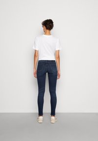 Jeans skinny in denim blu scuro con una texture liscia, abbinati a una maglietta bianca a maniche corte. Presentano tasche posteriori e un design aderente.