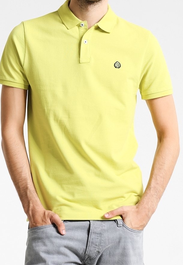 Homme portant un polo jaune à manches courtes avec un petit logo noir et blanc sur la poitrine, mains dans les poches, pantalon gris clair.