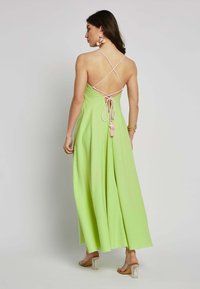 Langes, fließendes Kleid aus leicht grünem Stoff. Hat einen überkreuzten Rücken mit rosa Bändern und dekorativen Quasten. Durchsichtige Saumdetails, klare Absätze.