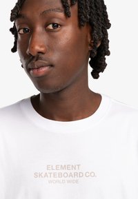 Camiseta blanca con "ELEMENT SKATEBOARD CO. WORLD WIDE" impreso en color beige claro. El modelo tiene el cabello corto y rizado de color oscuro y una expresión neutral.