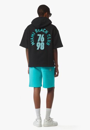 Schwarzer, übergroßer Hoodie mit "MIAMI BEACH CLUB" und Zahlen in Aquablau gedruckt, kombiniert mit Aquablaus-Shorts. Weiße Sneakers und Knöchelsocken vervollständigen das Outfit.